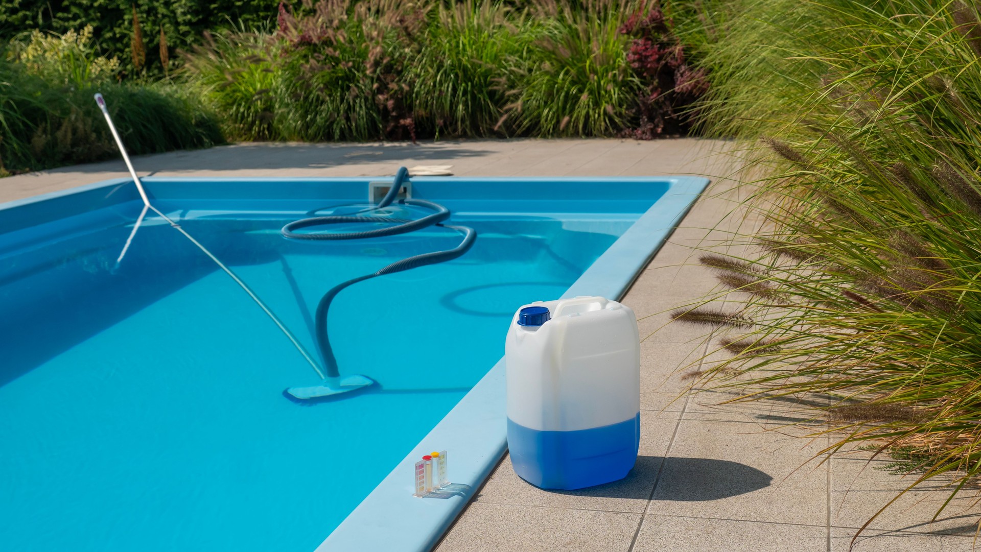 Pool cleaning hose and vacuum head with a large chemical container and water testing kit by the poolside, focusing on water quality and maintenance.
